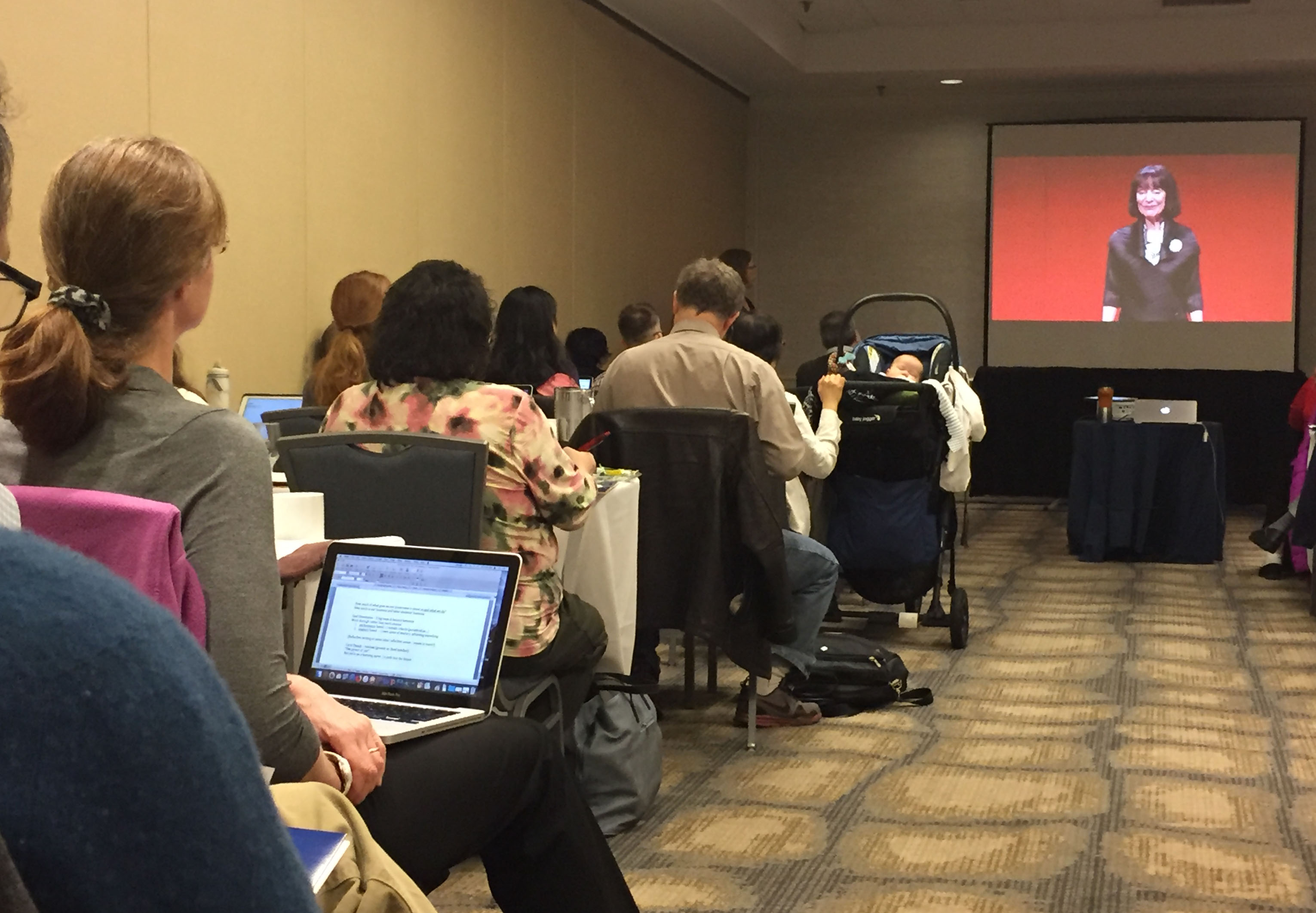 Attendees sit in rows watching a speaker on a video screen