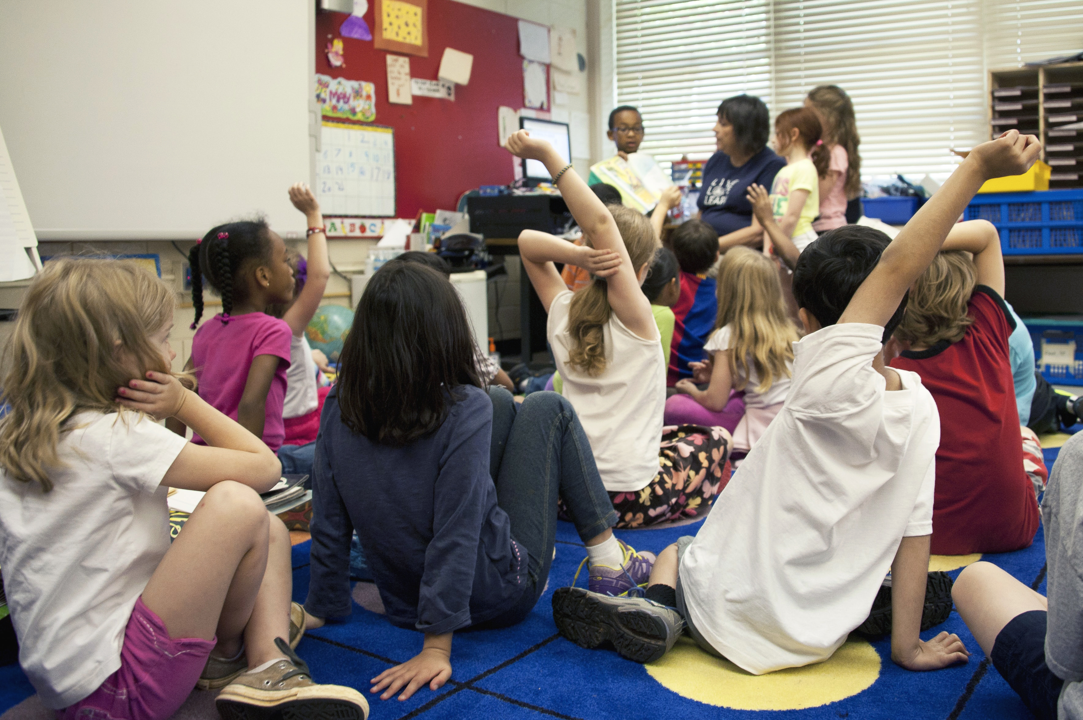 Early education classroom. Foreground includes children seated on the floor, backs to the camera, several with their hands raised. Background shows teacher surrounded by three students, one of whom is holding up a book for the class. 