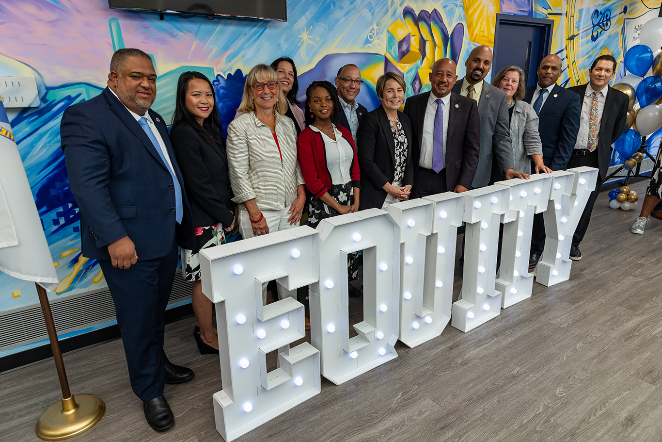 A group of twelve individuals, including Governor Maura Healey, Lt. Governor Kim Driscoll, Senate President Karen Spilka, Secretary of Education Patrick Tutwiler, and Commissioner of Higher Education Noe Ortega, are seen smiling and posing together. They are positioned in front of a vibrant mural and stand behind a large illuminated sign that prominently reads 'EQUITY.'